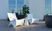 Two modern white chairs and a Ledge Lounger Autograph Standard Side Table with a potted orchid are arranged on a wooden patio deck, surrounded by green plants and large windows in the background.