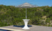 A black and white striped umbrella rests in the Ledge Lounger Autograph Umbrella Stand Ice Bin on a wooden deck by a pool, framed by green trees and a clear blue sky.