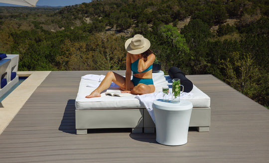 A woman in a blue bikini and sunhat relaxes on a lounge bed, reading by the Affinity Side Table from Ledge Lounger. Drinks rest atop the table as she enjoys nature and sunshine on a wooden deck surrounded by trees.