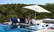 A woman relaxes on the blue Ledge Lounger Signature Sectional Middle with two-piece cushions beneath a large white umbrella by a modern pool, surrounded by loungers and lush greenery.