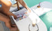 A person is stocking a PartyPad Swim-Up Bar by Ledge Lounger with canned drinks and ice beside the pool, perfect for poolside sipping. Nearby are sunglasses, a bottled drink, and snacks—setting the stage for a relaxing day by the water.