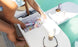 A person is stocking a PartyPad Swim-Up Bar by Ledge Lounger with canned drinks and ice beside the pool, perfect for poolside sipping. Nearby are sunglasses, a bottled drink, and snacks—setting the stage for a relaxing day by the water.