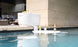 The Ledge Loungers PartyPad Swim-Up Bar, anchored by a toilet tank at the pools edge, invites poolside sipping with its floating white surface perfect for holding beer bottles and other beverages. In the background, modern patio seating enhances the relaxed atmosphere.