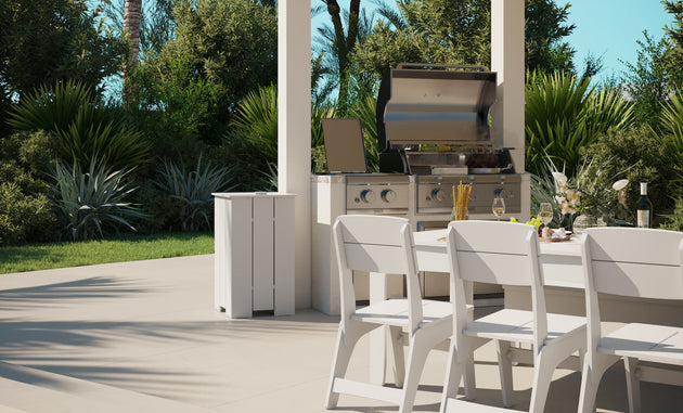 Outdoor dining is a delight with a white table and Ledge Loungers Mainstay Dining Side Chairs, perfectly set near a stainless steel grill. Plates, utensils, glasses, and vases adorn the scene amidst lush green foliage under a clear blue sky.