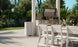 Outdoor dining is a delight with a white table and Ledge Loungers Mainstay Dining Side Chairs, perfectly set near a stainless steel grill. Plates, utensils, glasses, and vases adorn the scene amidst lush green foliage under a clear blue sky.