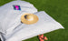A straw hat and a magazine rest on Ledge Loungers Laze Pillow with a subtle pattern, crafting an idyllic scene on the lush green grass. Nearby, orange sandals featuring red straps complete the outdoor comfort vibe.