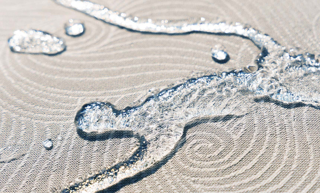 Close-up of clear water droplets on the beige spiral-patterned, waterproof fabric of the Ledge Lounger Laze Pillow, an outdoor comfort accessory. The droplets showcase the texture and water-repellent properties of this floating pool cushion.