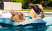 A blonde woman in a teal swimsuit lounges on a Ledge Lounger Laze Pillow floating in the pool, reading a magazine. She wears sunglasses and enjoys the outdoor comfort and bright sunlight surrounded by lush greenery and a stone wall.