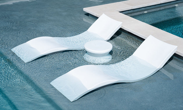 The Outdoor Oasis In-Pool Furniture Bundle by Ledge Lounger includes two Signature Chaise Loungers partially submerged and angled towards a small round table against a light blue tiled pool with visible ripples—the perfect poolside essential.