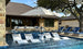 A luxurious backyard pool with white lounge chairs partially submerged in water features a Ledge Lounger Essential Fiberglass Umbrella - 9 Octagon Crank Auto Tilt, using marine-grade fabrics to shade a seating area. The stone house and trees create a serene, wind-resistant oasis for relaxation.