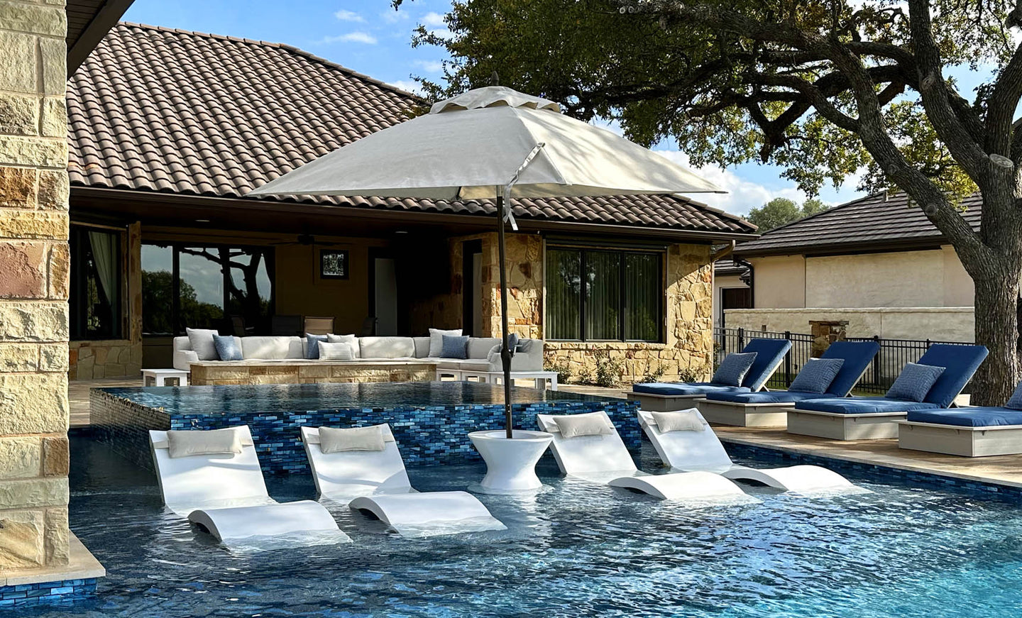 A stone house and large swimming pool with submerged loungers and blue sunbeds create a luxurious backyard. The area is shaded by the Essential Fiberglass Umbrella - 10 Square Pulley from Ledge Lounger, and is surrounded by trees under clear skies for a serene atmosphere.