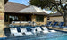 A stone house and large swimming pool with submerged loungers and blue sunbeds create a luxurious backyard. The area is shaded by the Essential Fiberglass Umbrella - 10 Square Pulley from Ledge Lounger, and is surrounded by trees under clear skies for a serene atmosphere.