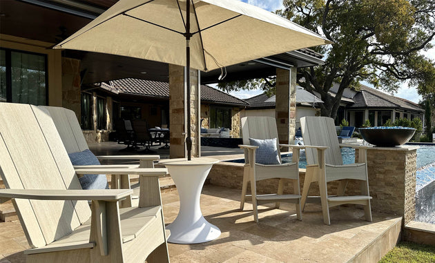 Two beige Adirondack chairs with cushions face a pool. Centered between them is a Ledge Lounger Essential Fiberglass Umbrella - 8.5 Rectangle Pulley, offering shade. A small white table sits nearby amidst stone structures and trees, creating a calm ambiance.