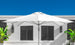 A 10 Square Pulley Essential Fiberglass Umbrella by Ledge Lounger stands open on a sunny day against a white building with dark shutters, framed by green plants and a clear blue sky, highlighting its durable fiberglass ribs and marine-grade fabrics.