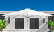 A 10 Square Pulley Essential Fiberglass Umbrella by Ledge Lounger stands open on a sunny day against a white building with dark shutters, framed by green plants and a clear blue sky, highlighting its durable fiberglass ribs and marine-grade fabrics.