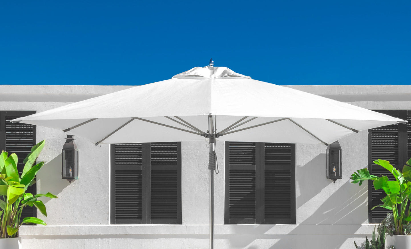 An 8.5 rectangle Essential Fiberglass Umbrella by Ledge Lounger, made from marine-grade fabrics, offers shade near a white building with dark shutters. Two wall-mounted lanterns and greenery are present on both sides under a clear blue sky.