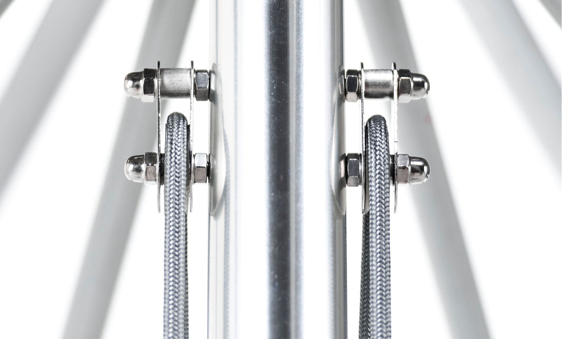 A close-up of a symmetrical metal structure with fiberglass ribs and vertical cables flanking a central pole mirrors the industrial aesthetic of an Essential Fiberglass Umbrella - 10 Square Pulley by Ledge Lounger against a bright white backdrop.
