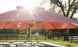 A 7.5 Essential Fiberglass Umbrella by Ledge Lounger provides shade over the pool and lounge chairs. Sunlight dapples through tree leaves, creating a warm ambiance in the grassy yard with a wooden fence. Its marine-grade fabric ensures durability all season long.
