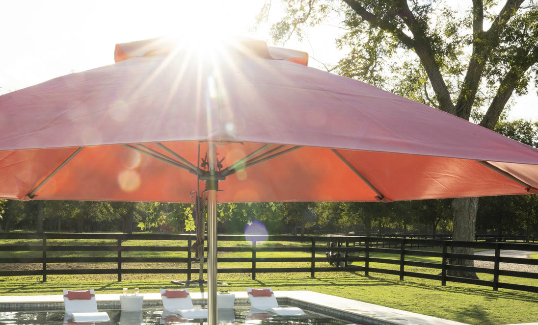 A 7.5 octagon Essential Fiberglass Umbrella by Ledge Lounger in red casts shade over a pool with submerged loungers. Sunlight filters through trees, creating a serene atmosphere while a wooden fence and lush greenery provide durability with marine-grade fabrics for a lasting oasis.