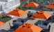 An outdoor seating area with round tables and chairs beneath large Ledge Lounger Essential Fiberglass Umbrellas - 7.5 Square Pulley on a paved patio. A white railing and stairs are visible on the left, against a rocky backdrop, combining style and durability with marine-grade fabrics.