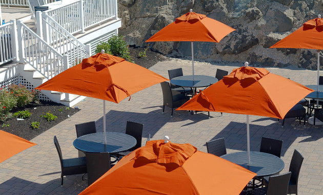 Circular tables with orange Ledge Lounger Essential Fiberglass Umbrellas featuring aluminum center poles are on a paved patio, surrounded by black chairs. A staircase and rock wall appear in the background.