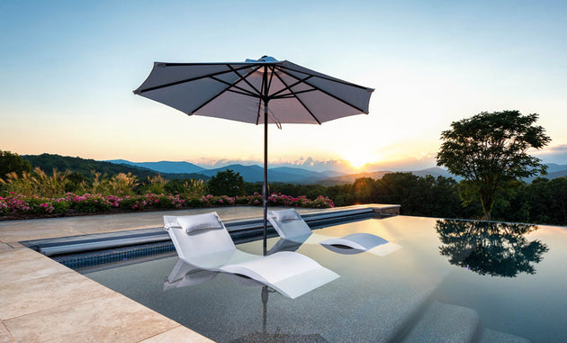 Two white lounge chairs are partly submerged in an infinity pool, shaded by Ledge Loungers Essential Fiberglass Umbrella - 7.5 Octagon Crank Auto Tilt, with a scenic mountain view and glowing sunset amidst surrounding greenery and flowers.