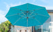 The Ultra Cantilever Umbrella, an 11 octagon by Ledge Lounger, stands open against a clear sky. Its sturdy metal frame casts shade over a welcoming patio with wicker furniture. Trees in the background enhance this serene setting further illuminated by an integrated outdoor light.