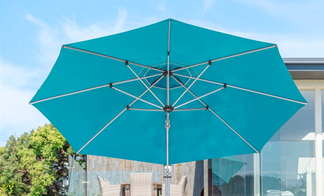 The teal Ultra Cantilever Umbrella by Ledge Lounger shades a stylish patio space under a clear blue sky. With outdoor chairs, a table visible beneath, and a rechargeable umbrella light adding evening charm, the scene is framed by trees and a building.