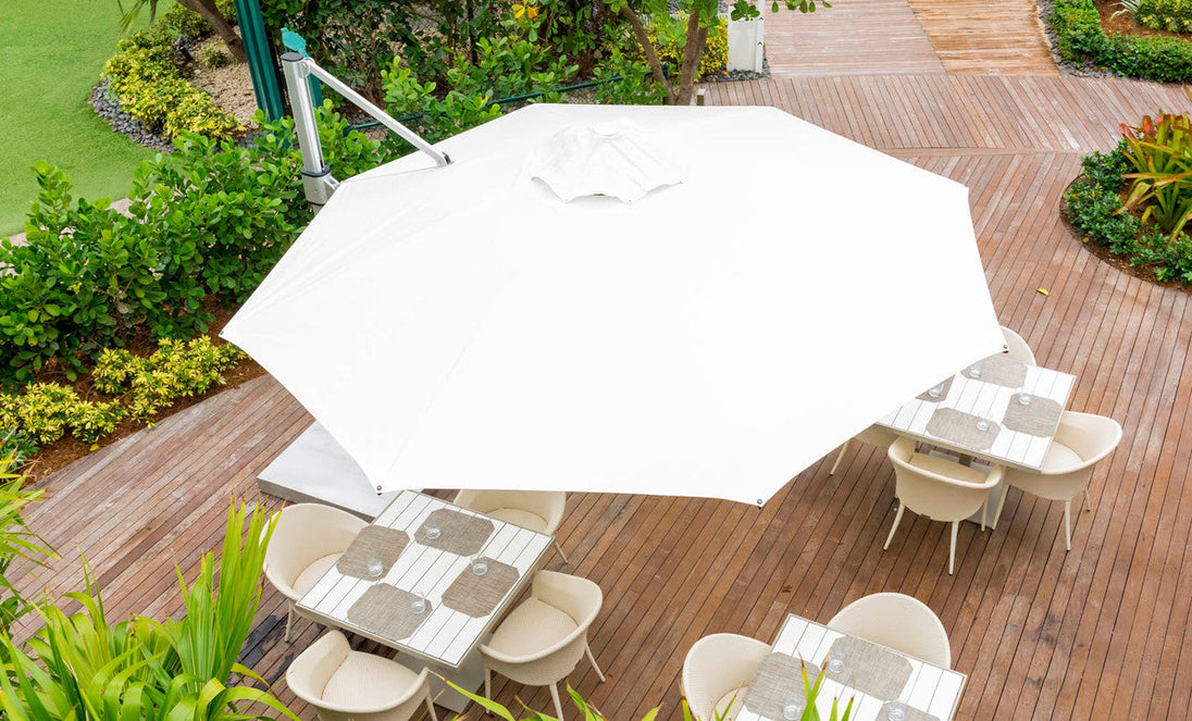 Aerial view of a patio with an outdoor dining space on a wooden deck, featuring the Ledge Lounger Ultra Cantilever Umbrella - 13 Octagon with rechargeable light illuminating tables set with placemats and encircled by white chairs, bordered by lush green foliage.
