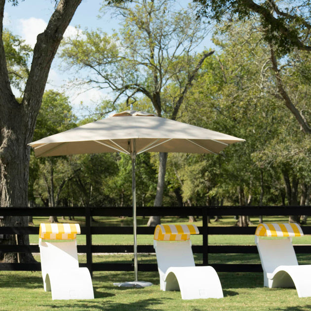 Three white lounge chairs with yellow-striped towels are shaded by a Ledge Lounger Premier Aluminum Umbrella - 7.5’ Octagon Pulley, crafted for durability with marine-grade fabrics, on grass framed by a black wooden fence under lush green trees and a clear blue sky.