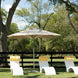 Three white lounge chairs with yellow-striped towels are shaded by a Ledge Lounger Premier Aluminum Umbrella - 7.5’ Octagon Pulley, crafted for durability with marine-grade fabrics, on grass framed by a black wooden fence under lush green trees and a clear blue sky.