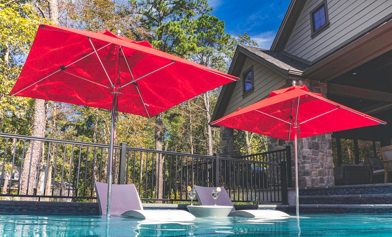 Two modern white lounge chairs partially submerged in a pool, each under a Ledge Lounger Premier Aluminum Umbrella - 7.5 Square Pulley. Set against a stone house and trees with a clear sky, the scene highlights marine-grade fabrics designed to withstand the elements.