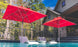 Two modern white lounge chairs partially submerged in a pool, each under a Ledge Lounger Premier Aluminum Umbrella - 7.5 Square Pulley. Set against a stone house and trees with a clear sky, the scene highlights marine-grade fabrics designed to withstand the elements.