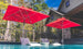 Two white lounge chairs partially submerged in a pool are shaded by Ledge Loungers wind-resistant Premier Aluminum Umbrella - 6.5 Square Pulley with marine-grade fabric. A stone house and surrounding trees enhance the serene outdoor atmosphere.