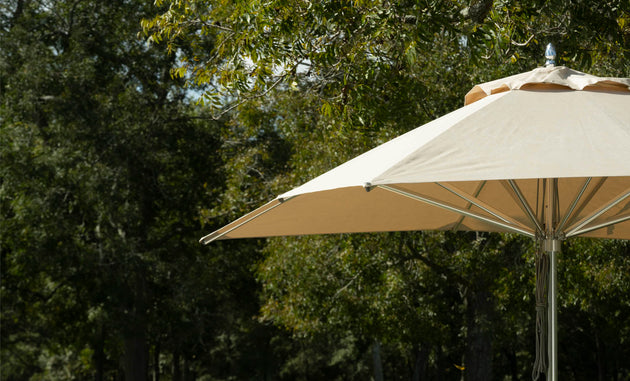 The Ledge Lounger Premier Aluminum Umbrella - 13 Octagon Pulley in beige is partially open, showcasing its wind-resistant design against lush green trees and blue sky, offering shade and a striking contrast to the natural landscape.