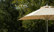 The Ledge Lounger Premier Aluminum Umbrella - 13 Octagon Pulley in beige is partially open, showcasing its wind-resistant design against lush green trees and blue sky, offering shade and a striking contrast to the natural landscape.