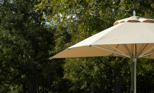 A Premier Aluminum Umbrella - 9 Octagon Pulley by Ledge Lounger offers shade beneath lush trees on a sunny day. Made with marine-grade fabrics, the partially open large beige umbrella combines style and wind resistance against the backdrop of dense foliage and dappled sunlight.