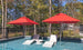 Two white lounge chairs and a small round table sit partially submerged in a shallow pool, shaded by two wind-resistant Ledge Lounger Premier Aluminum Umbrellas made from marine-grade fabrics. A black fence surrounds the pool, with trees in the background.