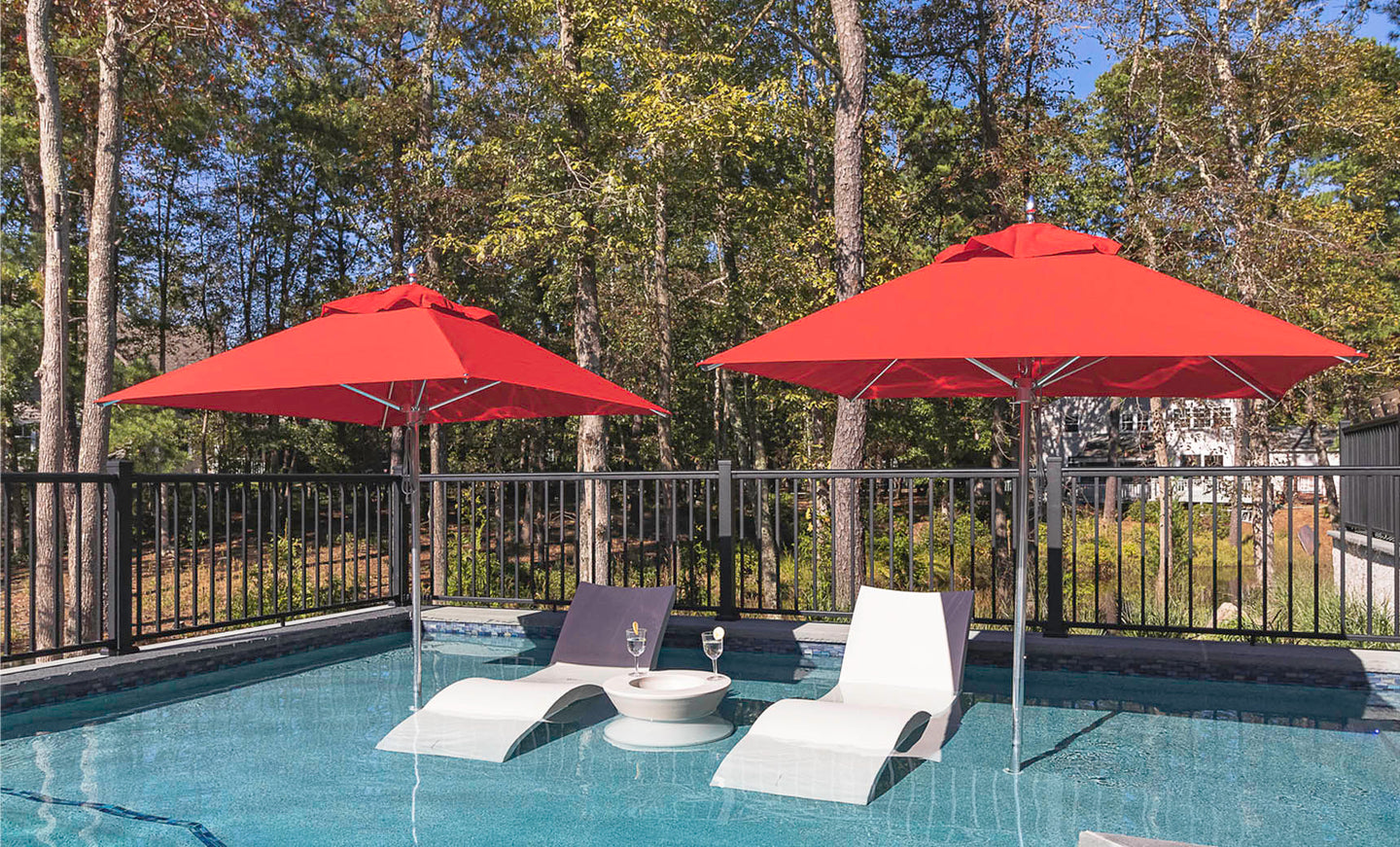 Two white lounge chairs and a small round table with a wine glass are set in a shallow pool area under two Ledge Lounger Premier Aluminum Umbrellas - 8.5 Rectangle Pulley, made with marine-grade fabrics. Black fencing and tall trees provide a serene backdrop.