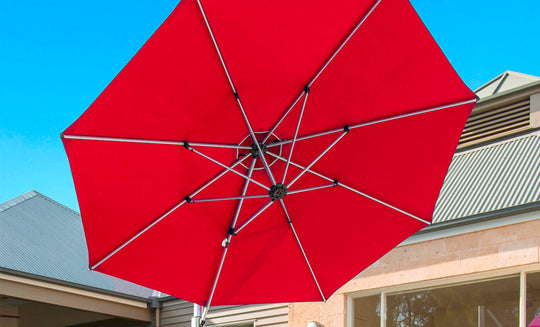 The Ultra Cantilever Umbrella by Ledge Lounger, a 13 red octagon, extends under a clear blue sky with a chic rechargeable umbrella light. Its mounted on a metal frame against the backdrop of a house with a slanted roof.