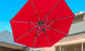 The Ultra Cantilever Umbrella by Ledge Lounger, a 13 red octagon, extends under a clear blue sky with a chic rechargeable umbrella light. Its mounted on a metal frame against the backdrop of a house with a slanted roof.