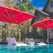 Two white lounge chairs with attached tables sit in a clear pool under the shade of two 7.5 Premier Aluminum Umbrellas with marine-grade fabric by Ledge Lounger. A stone house with green siding and black railing is surrounded by trees, all under a clear blue sky.