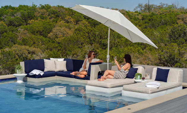 Two women relax by a pool on a chic outdoor sectional. One lounges on a navy sofa with a rustproof frame, the other enjoys the comfort of a striped chaise under Ledge Loungers 6.5 Square Signature 360° Tilt Umbrella, surrounded by greenery as they savor the sunny day.