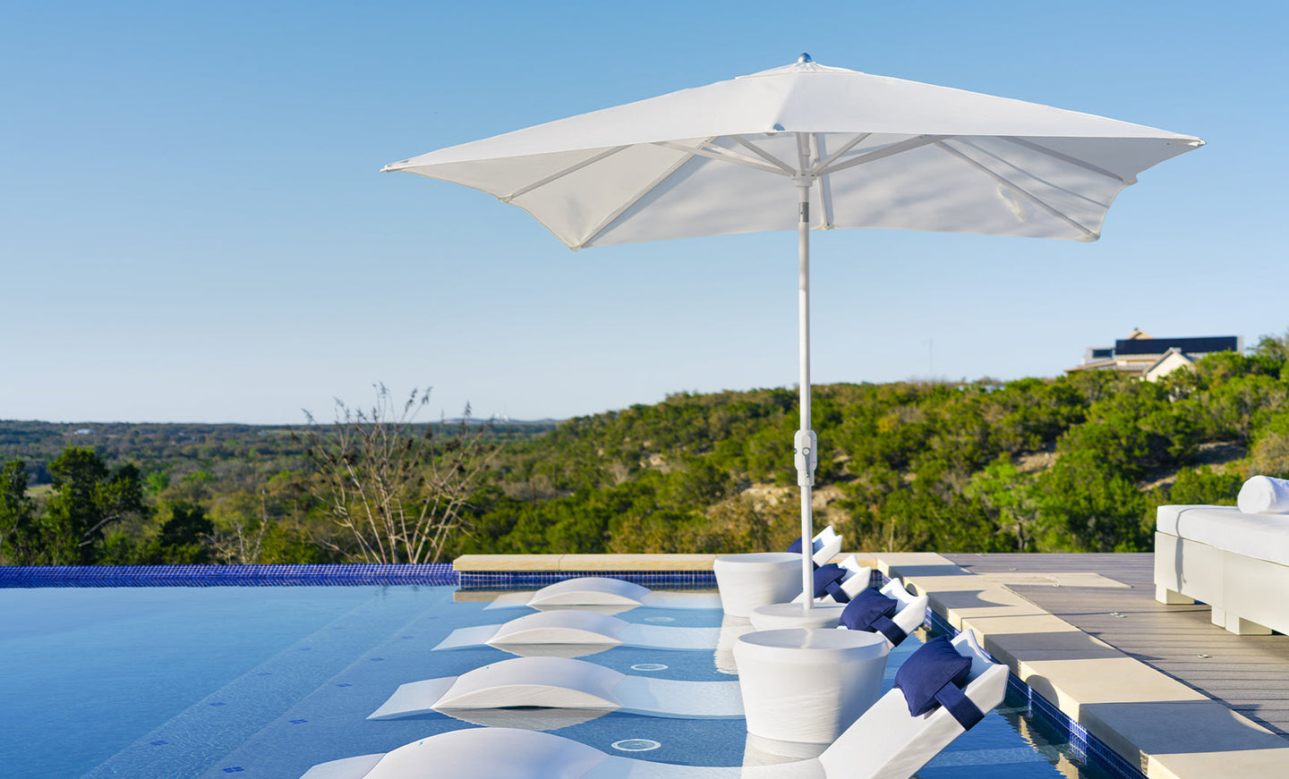 A tranquil outdoor scene showcases Ledge Loungers rustproof white lounge chairs with blue cushions, partially submerged in an infinity pool. A 6.5 Square Signature 360° Tilt Umbrella offers shade, set against a vast tree-covered landscape under a clear blue sky.