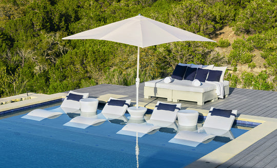 A modern poolside oasis includes white lounge chairs partially submerged in water, perfectly shaded by a Ledge Lounger 7.5 Octagon Signature 360° Tilt Umbrella. A double daybed with blue and white cushions sits on a wooden deck amidst lush greenery.