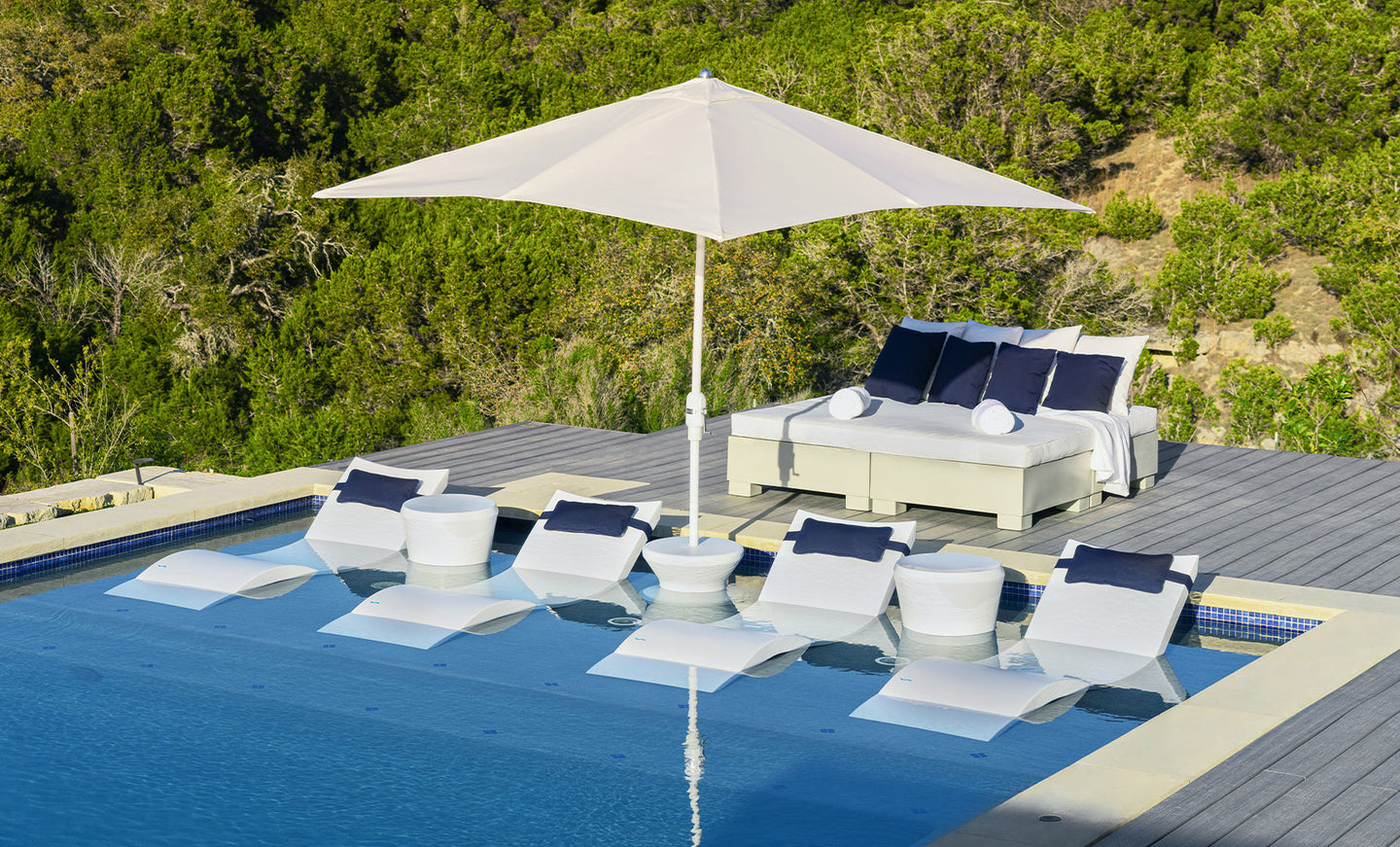 An outdoor pool scene features six white lounge chairs partially submerged, shaded by a 6.5 Square Signature 360° Tilt Umbrella by Ledge Lounger. A daybed with blue and white pillows sits on the deck, surrounded by lush greenery and a hillside backdrop.