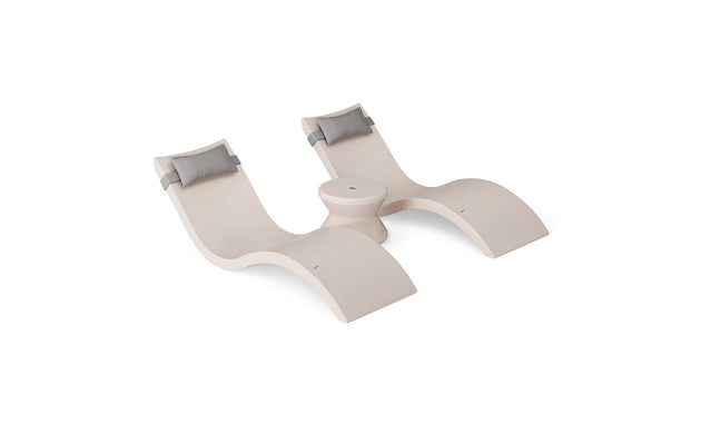 The Ledge Lounger Signature Chaise Headrest Pillow and Side Table In-Pool Set features two modern beige chaise loungers with gray headrest pillows, connected by a round center table, all styled for poolside chic against a white background.