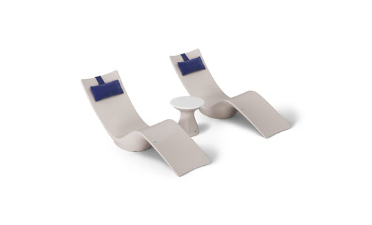 Two beige chaise loungers with blue headrests and a small, round white table create a minimalist, stylish setup ideal for poolside relaxation. Enjoy this aesthetic with the Resort Luxury Outdoor Paradise In-Pool Furniture Bundle from Ledge Lounger.