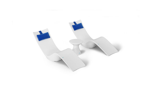 The Resort Luxury Outdoor Paradise In-Pool Furniture Bundle by Ledge Lounger includes two modern white chaise loungers with blue headrests and a small round white table, all featuring a sleek curved design, perfect for poolside relaxation. The set is showcased against a plain white background.