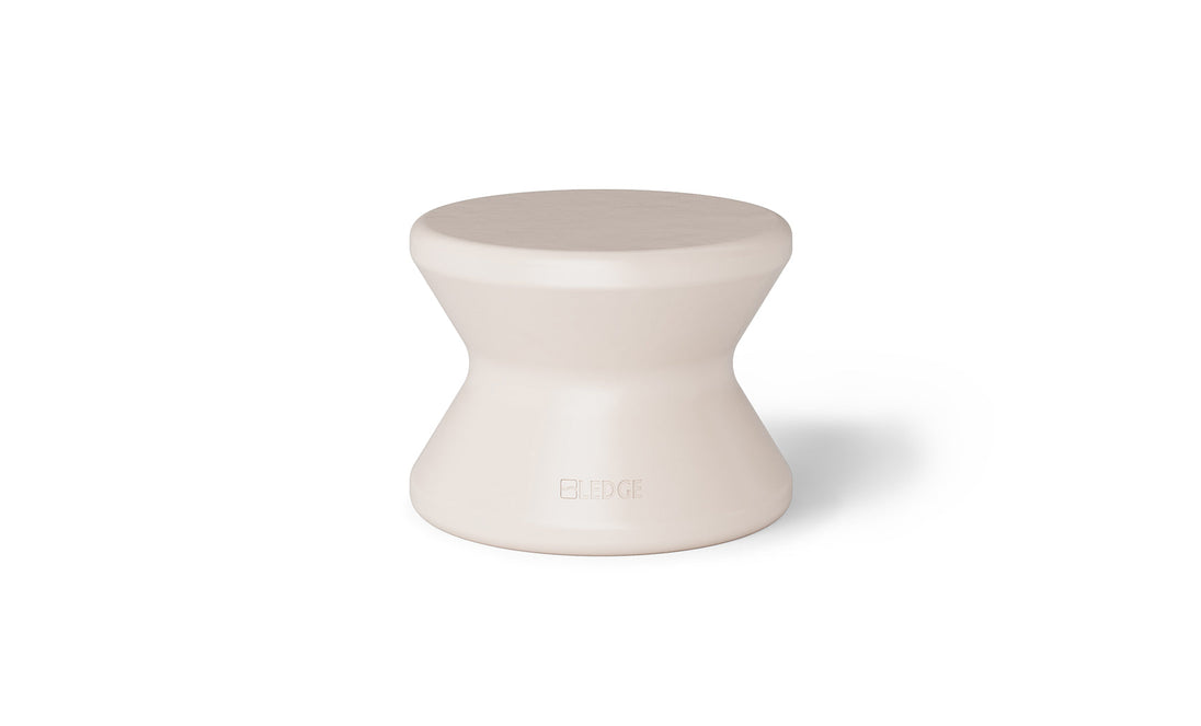 The Signature Standard Side Table by Ledge Lounger is a small, hourglass-shaped, light beige stool with a smooth surface for in-pool use on sun shelves. It features the word EDGE inscribed near the bottom, adding elegance and functionality.